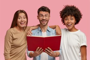 Indoor Shot of Cheerful Handsome Man and His Two Sisters Embrace and Stand Closely, Read Book, Have Happy Expression, like Learning Something New, Isolated over Pink Background. Teamwork Concept