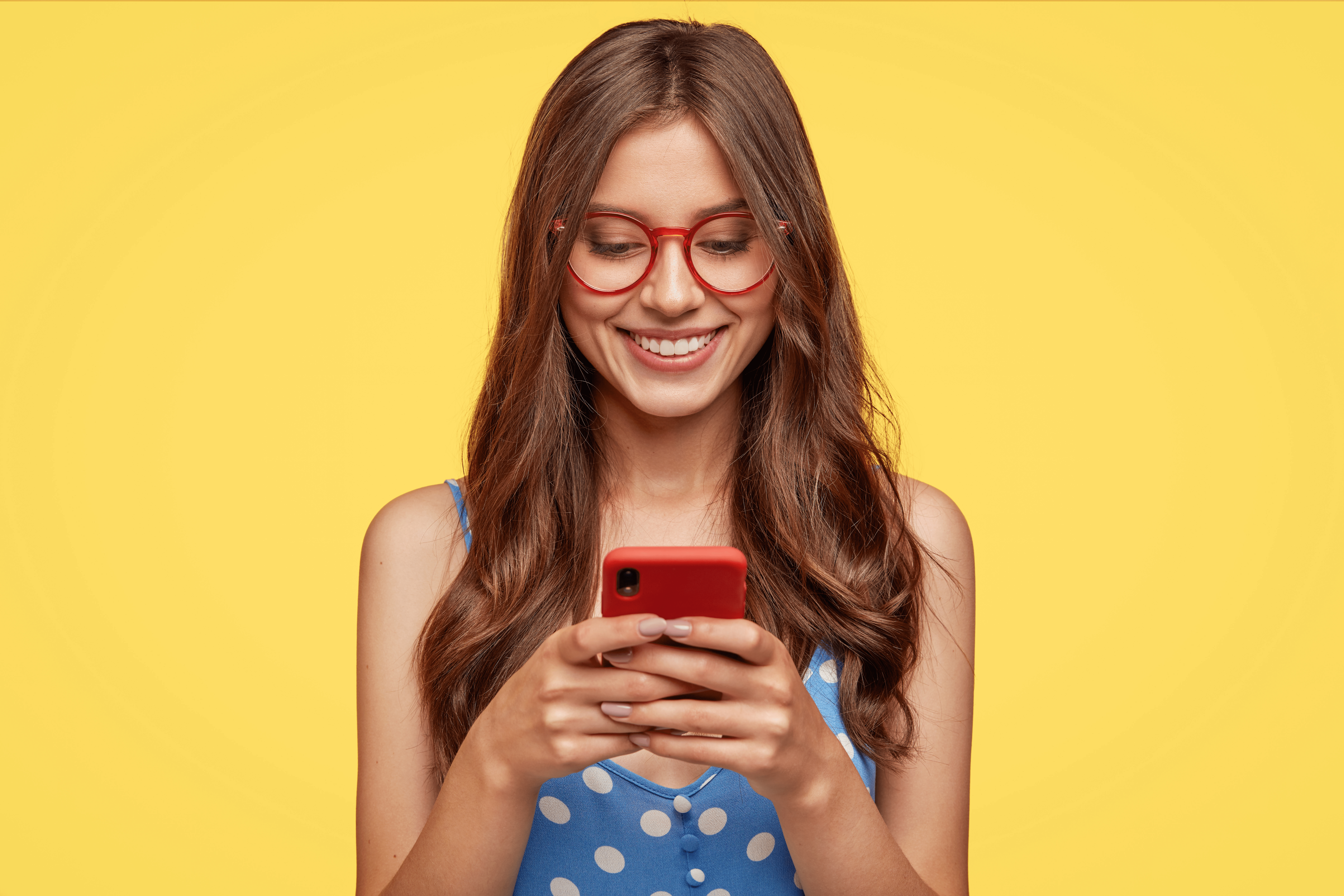 Content Teenager with Long Hair, Holds Modern Cell Phone, Scrolls through Social Networks, Has Cheerful Expression, Wears Spectacles and Casual Dress, Isolated over Yellow Background, Recieves Message