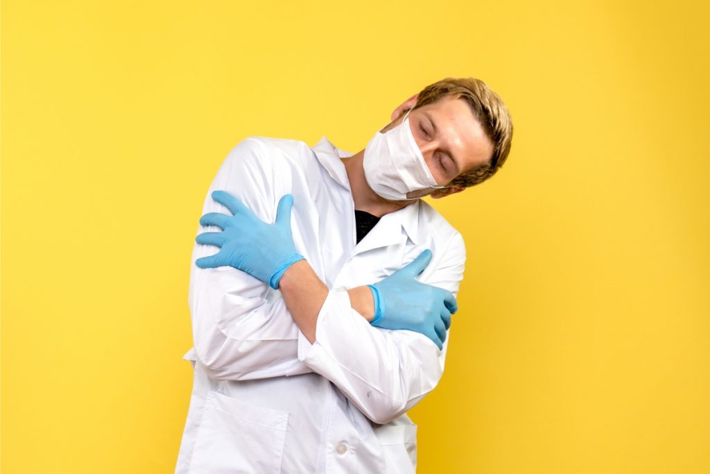 image of a male, white doctor wearing blue rubber gloves, a white mask and a white coat, hugging himself with smiling eyes against a bright yellow background