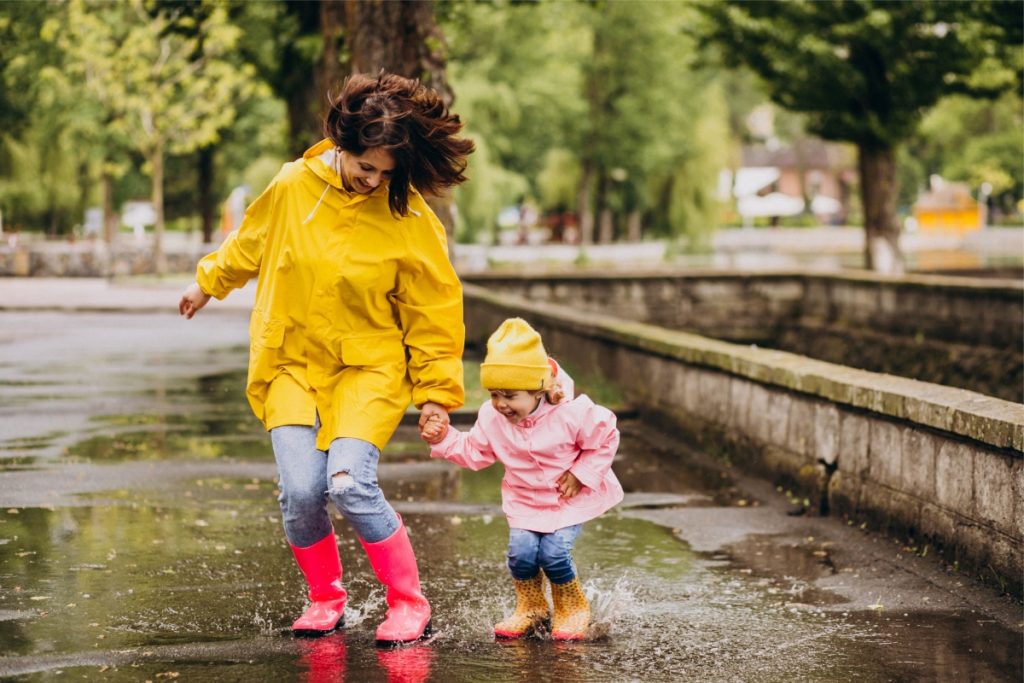 brunette white woman wearing a yellow coat and red rain boots jumps in a puddle with her daughter who wears a yellow hat and pink coat